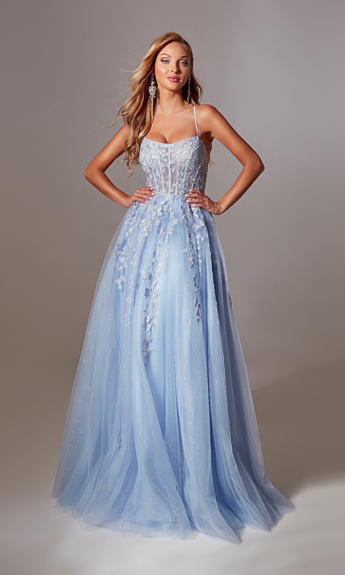 Model wears the Aerie Couture 266 Lace-Up Long Prom Ball Gown—a light blue, floor-length embroidered lace dress with floral appliqués, thin straps, sheer tulle overlay, and lace-up open back—posing hands on hips against a neutral backdrop.